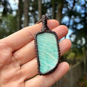 Pode incluir: Um pingente retangular com uma pedra azul-esverdeada clara com um padr&atilde;o &uacute;nico, envolto em macram&ecirc; tecido marrom escuro. O pingente &eacute; suspenso por um cord&atilde;o tecido correspondente, mostrando uma est&eacute;tica natural e terrosa. O fundo &eacute; uma vista desfocada de &aacute;rvores.