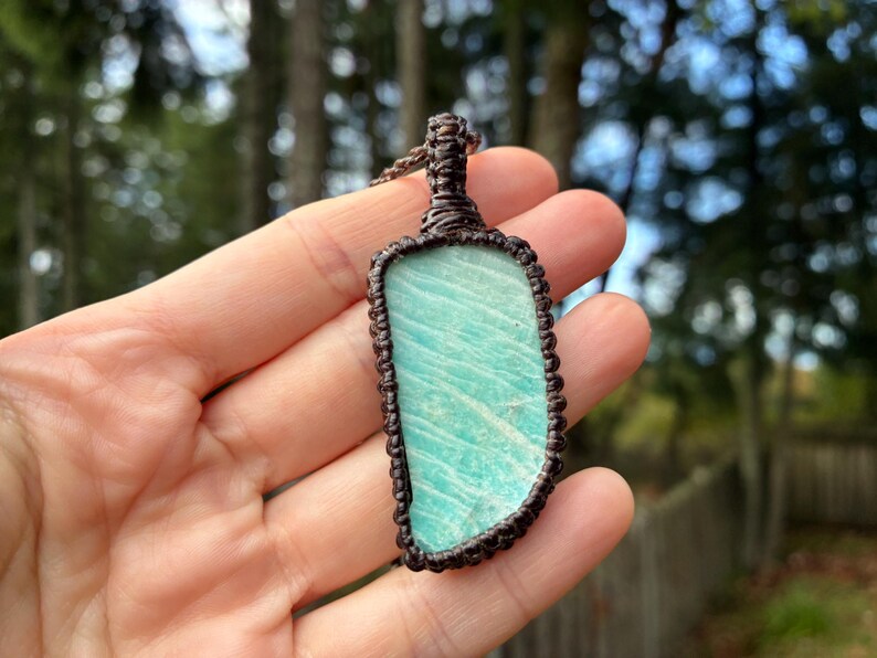 Pode incluir: Um pingente retangular com uma pedra azul-esverdeada clara com um padr&atilde;o &uacute;nico, envolto em macram&ecirc; tecido marrom escuro. O pingente &eacute; suspenso por um cord&atilde;o tecido correspondente, mostrando uma est&eacute;tica natural e terrosa. O fundo &eacute; uma vista desfocada de &aacute;rvores.