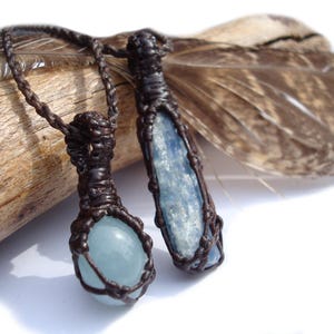 May include: Two necklaces with blue gemstone pendants wrapped in brown cord. One pendant is a round, light blue gemstone, and the other is a long, pointed, light blue gemstone.