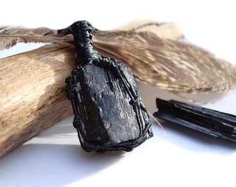 May include: A black tourmaline crystal pendant wrapped in black wire. The pendant is displayed with a piece of driftwood and a feather. The crystal is a dark, opaque color, and the wire wrapping adds a decorative element.