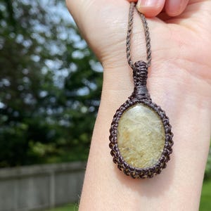 May include: A handmade macrame necklace featuring an oval-shaped, light-yellow gemstone pendant. The pendant is encased in a brown macrame weave and suspended from a matching brown cord. The background is a blurred outdoor scene with green foliage.