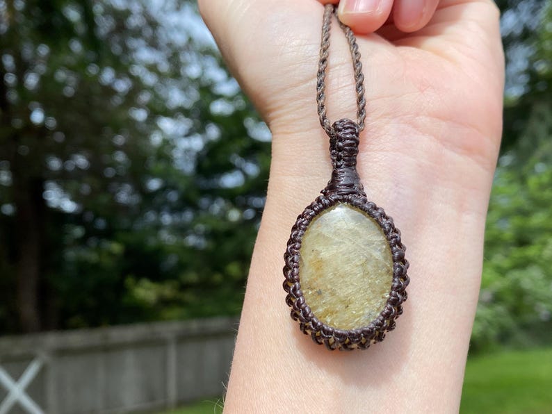 May include: A handmade macrame necklace featuring an oval-shaped, light-yellow gemstone pendant. The pendant is encased in a brown macrame weave and suspended from a matching brown cord. The background is a blurred outdoor scene with green foliage.
