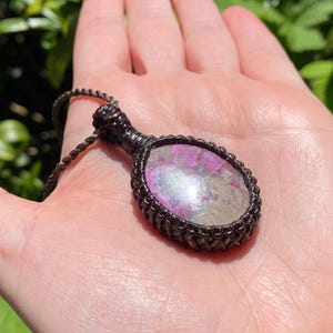 May include: A handcrafted pendant necklace featuring a large, oval-shaped stone with pink and iridescent hues, encased in a dark brown macrame setting. The necklace is displayed on a hand, with a green leafy background.