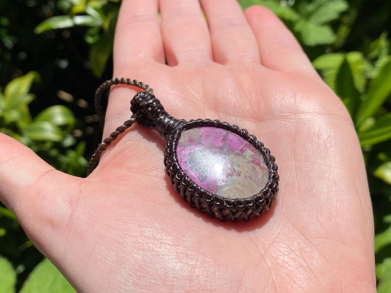 May include: A handcrafted pendant necklace featuring a large, oval-shaped stone with pink and iridescent hues, encased in a dark brown macrame setting. The necklace is displayed on a hand, with a green leafy background.