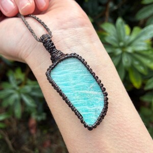 Pode incluir: Um colar com pingente artesanal com uma pedra preciosa retangular azul clara. A pedra &eacute; emoldurada em um design de macram&ecirc; marrom escuro e suspensa por um cord&atilde;o correspondente. O colar &eacute; exibido em um fundo verde natural. A pedra tem cerca de 5 cm de comprimento.