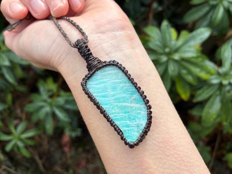 Pode incluir: Um colar com pingente artesanal com uma pedra preciosa retangular azul clara. A pedra &eacute; emoldurada em um design de macram&ecirc; marrom escuro e suspensa por um cord&atilde;o correspondente. O colar &eacute; exibido em um fundo verde natural. A pedra tem cerca de 5 cm de comprimento.