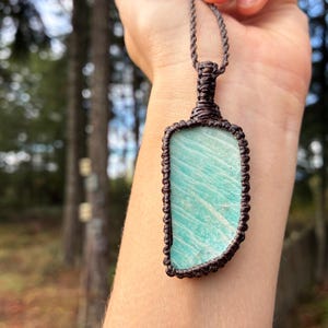 Pode incluir: Um colar com pingente retangular com uma pedra azul clara e uma borda tecida marrom. O pingente &eacute; suspenso por um cord&atilde;o marrom correspondente. O fundo est&aacute; desfocado, com &aacute;rvores vis&iacute;veis.