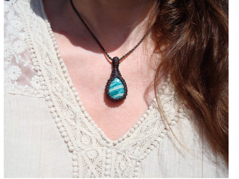 May include: A handcrafted macrame necklace featuring a teardrop-shaped pendant. The pendant showcases a vibrant blue-green stone with white veining, encased in a dark brown macrame weave. The necklace is worn against a cream-colored blouse.