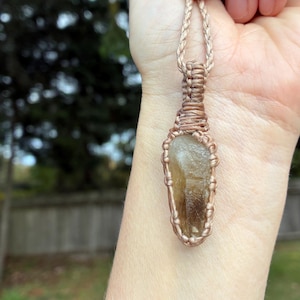 May include: A handcrafted pendant necklace featuring a light brown crystal encased in a copper-colored wire wrap. The crystal is suspended from a woven cord. The background shows a blurred outdoor scene with trees and a fence.
