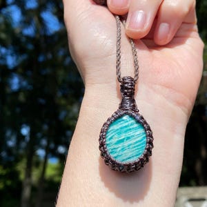 May include: A handcrafted pendant necklace featuring a teardrop-shaped amazonite stone. The stone is a vibrant turquoise color with white veining, set in a dark brown macrame design. The necklace has a matching brown cord.