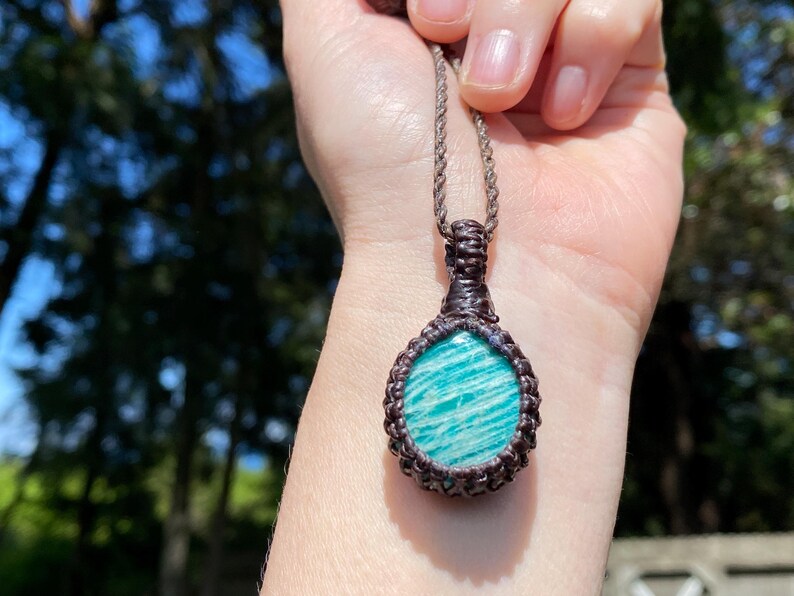 May include: A handcrafted pendant necklace featuring a teardrop-shaped amazonite stone. The stone is a vibrant turquoise color with white veining, set in a dark brown macrame design. The necklace has a matching brown cord.