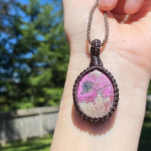 May include: A handcrafted pendant necklace featuring a teardrop-shaped stone with pink and gray hues, encased in a dark brown macrame setting. The pendant hangs from a matching brown cord, showcasing a natural, earthy aesthetic.