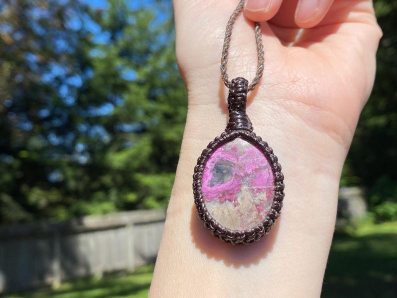 May include: A handcrafted pendant necklace featuring a teardrop-shaped stone with pink and gray hues, encased in a dark brown macrame setting. The pendant hangs from a matching brown cord, showcasing a natural, earthy aesthetic.