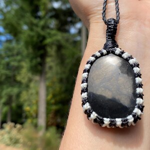 K&ouml;nnte beinhalten: Eine handgefertigte Anh&auml;ngerkette mit einem ovalen, polierten Stein in einer Makramee-Fassung. Das Makramee-Design verwendet schwarze und wei&szlig;e Kordel, und die Halskette ist schwarz. Der Stein hat eine dunkle, reflektierende Oberfl&auml;che.