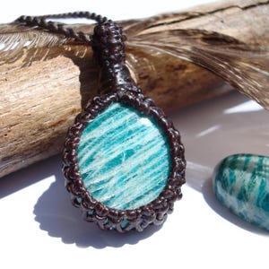 May include: A handcrafted pendant necklace featuring a polished, oval-shaped amazonite stone with a blue-green hue. The stone is encased in a dark brown macrame setting and hangs from a matching cord. A similar amazonite stone is visible to the right.
