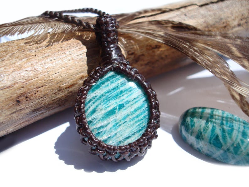 May include: A handcrafted pendant necklace featuring a polished, oval-shaped amazonite stone with a blue-green hue. The stone is encased in a dark brown macrame setting and hangs from a matching cord. A similar amazonite stone is visible to the right.