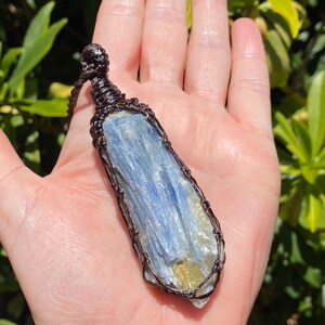 May include: A blue kyanite pendant with a brown wire wrap. The kyanite has a layered appearance with shades of blue and yellow. The pendant is suspended from a brown cord. The background is a hand and green foliage.