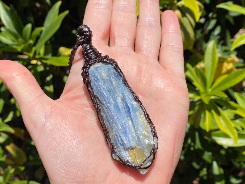 May include: A blue kyanite pendant with a brown wire wrap. The kyanite has a layered appearance with shades of blue and yellow. The pendant is suspended from a brown cord. The background is a hand and green foliage.