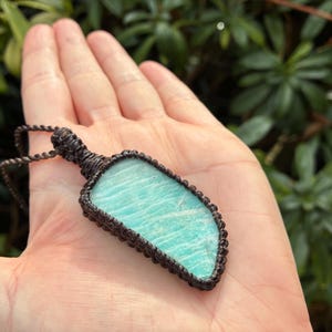 Pode incluir: Pingente artesanal com uma pedra preciosa azul clara polida em forma irregular. A pedra &eacute; emoldurada em uma moldura de macram&ecirc; marrom escuro, suspensa por um cord&atilde;o correspondente. O pingente &eacute; segurado em uma m&atilde;o aberta contra um fundo de folhagem verde.