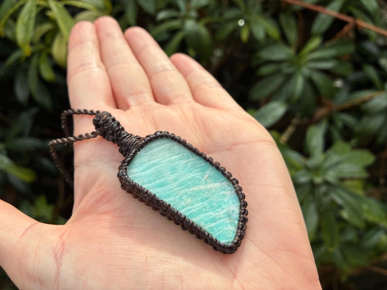 Pode incluir: Pingente artesanal com uma pedra preciosa azul clara polida em forma irregular. A pedra &eacute; emoldurada em uma moldura de macram&ecirc; marrom escuro, suspensa por um cord&atilde;o correspondente. O pingente &eacute; segurado em uma m&atilde;o aberta contra um fundo de folhagem verde.