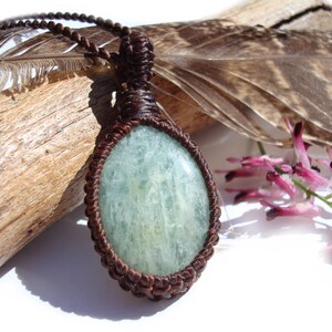 May include: A handcrafted macrame necklace featuring a light blue oval gemstone pendant. The pendant is encased in a brown woven macrame setting and hangs from a matching cord. The necklace is displayed on a piece of driftwood with a feather and pink flowers.