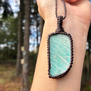 Pode incluir: Um pingente retangular com uma pedra azul clara com uma borda de macram&ecirc; marrom e um cord&atilde;o correspondente. A pedra tem um padr&atilde;o &uacute;nico de linhas azuis claras e escuras. O pingente &eacute; uma joia.