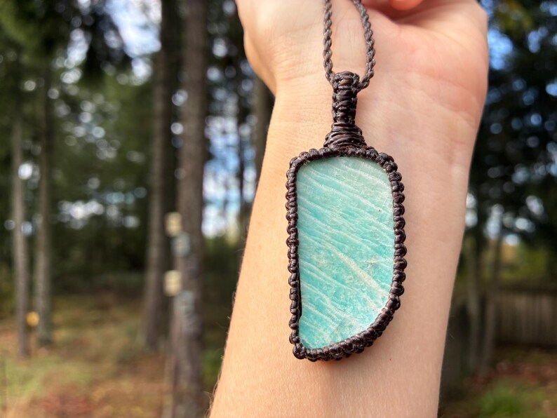 Pode incluir: Um pingente retangular com uma pedra azul clara com uma borda de macram&ecirc; marrom e um cord&atilde;o correspondente. A pedra tem um padr&atilde;o &uacute;nico de linhas azuis claras e escuras. O pingente &eacute; uma joia.