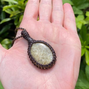 May include: A brown macrame necklace with a golden-colored oval stone pendant. The pendant is held in an open hand against a backdrop of green foliage. The necklace cord is also brown.