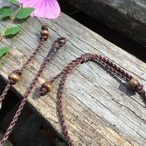 May include: Close-up of several brown braided cords with wooden beads, possibly for jewelry or crafts. The cords are arranged on a weathered wooden surface, with a pink flower and green leaves in the background. The cords have a natural, earthy aesthetic.