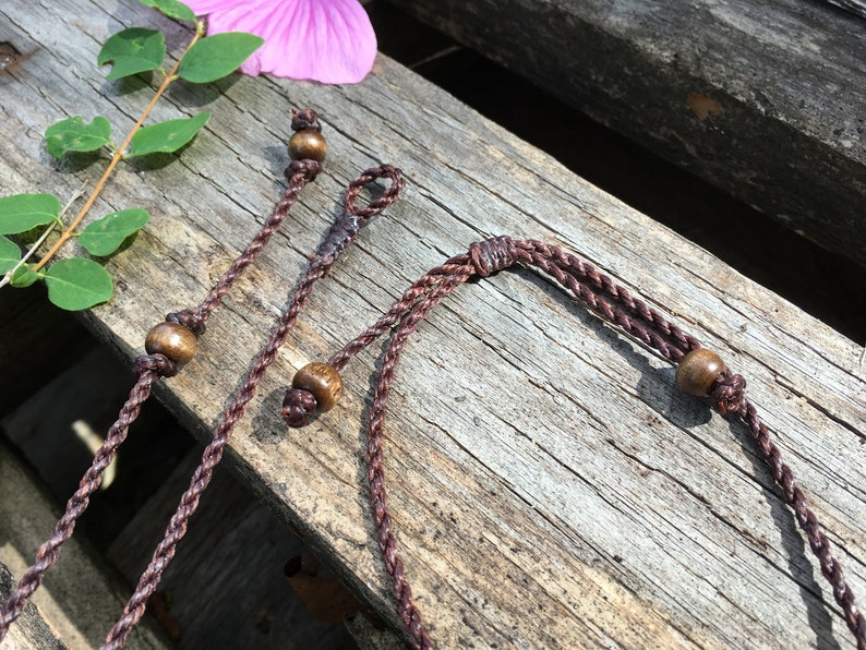 May include: Close-up of several brown braided cords with wooden beads, possibly for jewelry or crafts. The cords are arranged on a weathered wooden surface, with a pink flower and green leaves in the background. The cords have a natural, earthy aesthetic.