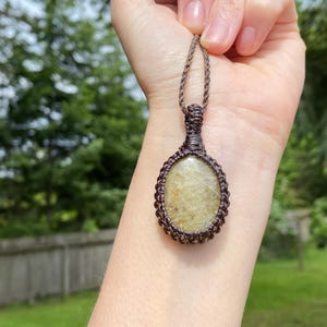 May include: A handcrafted macrame pendant necklace featuring an oval-shaped, light-colored stone. The stone is encased in a dark brown macrame weave, with a matching cord for wearing. The background is a blurred view of green foliage.
