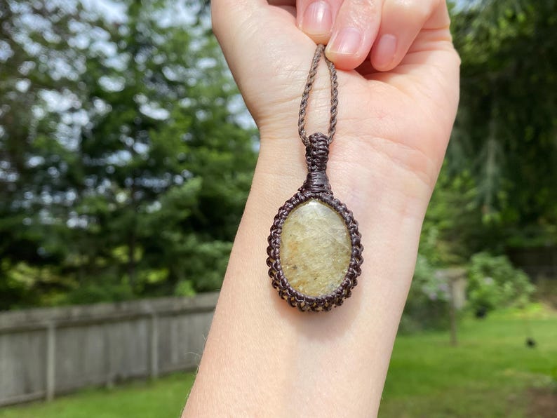 May include: A handcrafted macrame pendant necklace featuring an oval-shaped, light-colored stone. The stone is encased in a dark brown macrame weave, with a matching cord for wearing. The background is a blurred view of green foliage.