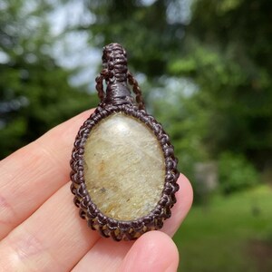 May include: A handcrafted pendant featuring a light-colored, oval-shaped stone encased in a dark brown macrame design. The pendant is suspended from a matching macrame cord, showcasing a natural, earthy aesthetic.