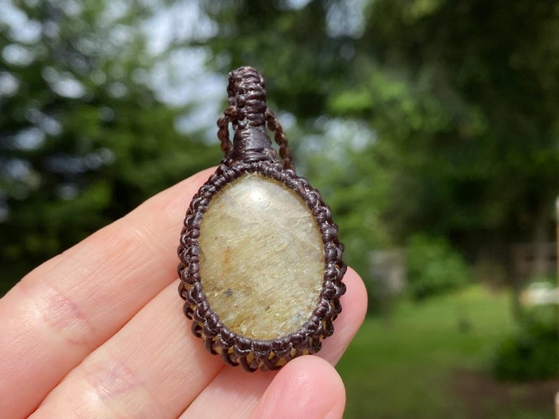 May include: A handcrafted pendant featuring a light-colored, oval-shaped stone encased in a dark brown macrame design. The pendant is suspended from a matching macrame cord, showcasing a natural, earthy aesthetic.