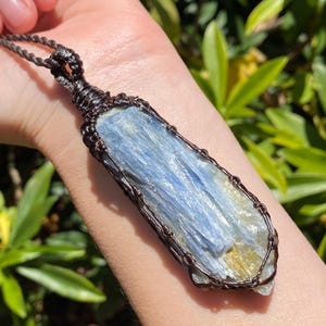 May include: A blue kyanite pendant necklace. The rectangular crystal is wrapped in dark brown wire and suspended from a matching cord. The kyanite has a layered appearance with shades of blue and a hint of yellow.
