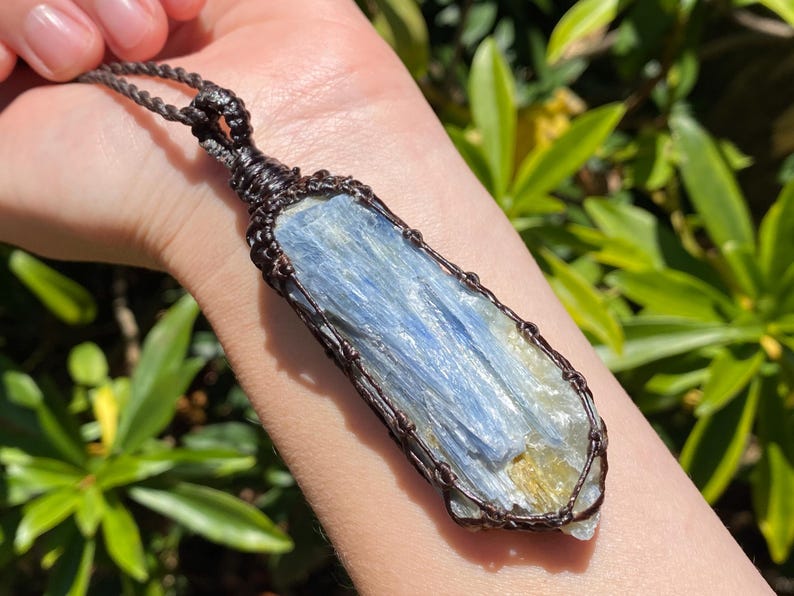 May include: A blue kyanite pendant necklace. The rectangular crystal is wrapped in dark brown wire and suspended from a matching cord. The kyanite has a layered appearance with shades of blue and a hint of yellow.