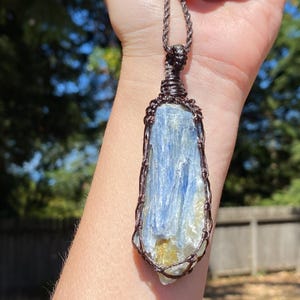 May include: A blue kyanite pendant, wire-wrapped in a copper-colored metal, hangs from a brown cord. The kyanite crystal has a layered appearance with shades of blue and a hint of yellow. The pendant is displayed against a blurred background of green trees.