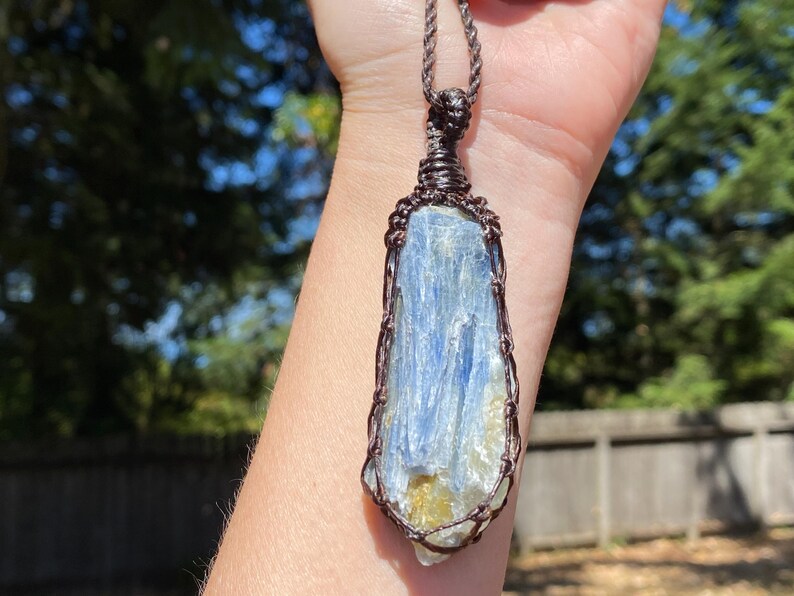 May include: A blue kyanite pendant, wire-wrapped in a copper-colored metal, hangs from a brown cord. The kyanite crystal has a layered appearance with shades of blue and a hint of yellow. The pendant is displayed against a blurred background of green trees.