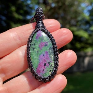 May include: A handcrafted pendant necklace featuring a large, oval-shaped gemstone. The stone displays a vibrant mix of green and magenta hues, encased in a black woven macrame setting. The necklace is displayed against a blurred outdoor background.