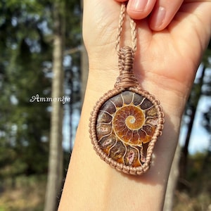 Pode incluir: Pingente artesanal com um fóssil de amonite polido, apresentando um padrão espiral em tons de castanho e dourado. O fóssil é emoldurado em macramé com um cordão tecido. A palavra "Amonite" está escrita em itálico.