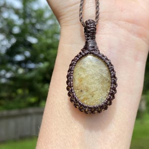 May include: A handmade macrame necklace featuring an oval-shaped, light-yellow gemstone pendant. The pendant is encased in a dark brown macrame weave and hangs from a matching cord. The background is a blurred outdoor scene with green foliage.