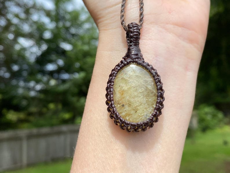 May include: A handmade macrame necklace featuring an oval-shaped, light-yellow gemstone pendant. The pendant is encased in a dark brown macrame weave and hangs from a matching cord. The background is a blurred outdoor scene with green foliage.
