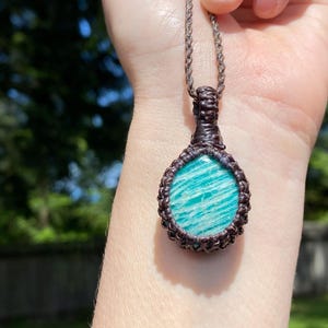 May include: A handcrafted macrame necklace featuring a round, light blue gemstone pendant with white veining. The pendant is encased in a dark brown macrame weave and hangs from a matching cord. The background is a blurred view of green foliage.