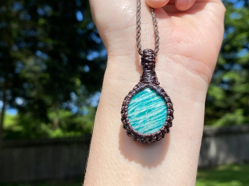 May include: A handcrafted macrame necklace featuring a round, light blue gemstone pendant with white veining. The pendant is encased in a dark brown macrame weave and hangs from a matching cord. The background is a blurred view of green foliage.