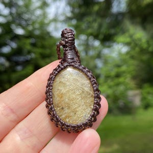 May include: Oval-shaped pendant with a light brown stone, encased in a macrame weave. The pendant is suspended from a matching cord. The stone has a golden hue with subtle variations in color.
