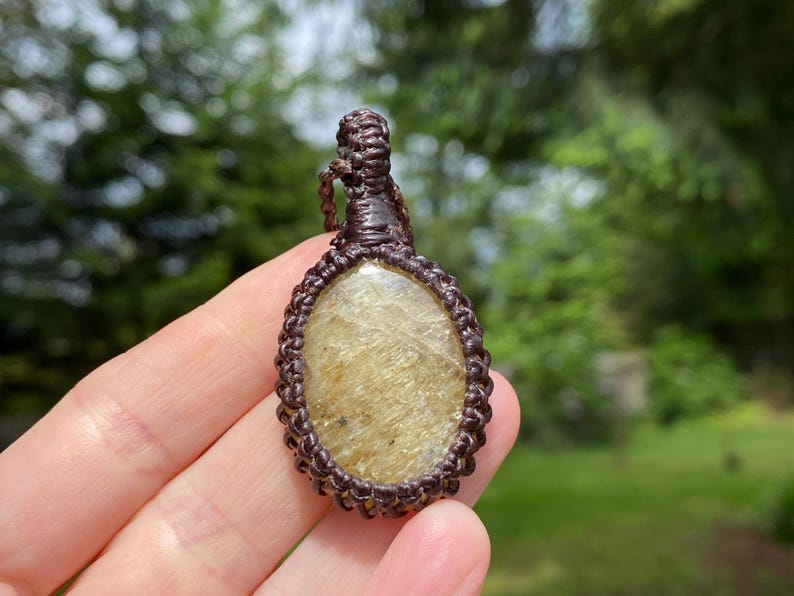 May include: Oval-shaped pendant with a light brown stone, encased in a macrame weave. The pendant is suspended from a matching cord. The stone has a golden hue with subtle variations in color.