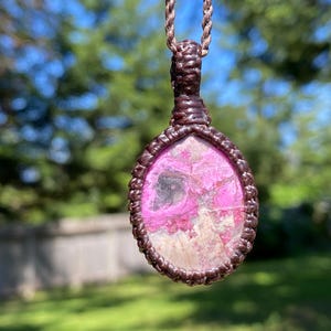 May include: A handcrafted pendant necklace featuring a pink and white stone set in a brown macrame design. The pendant is oval-shaped and suspended from a matching brown cord, creating a natural and earthy aesthetic.