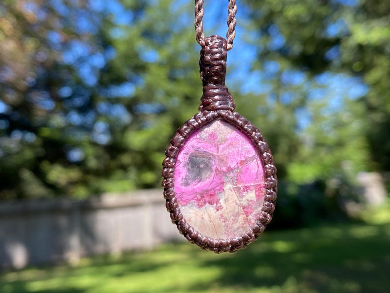 May include: A handcrafted pendant necklace featuring a pink and white stone set in a brown macrame design. The pendant is oval-shaped and suspended from a matching brown cord, creating a natural and earthy aesthetic.