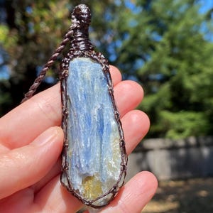 May include: A blue kyanite pendant necklace. The raw, elongated crystal is wrapped in brown wire and suspended from a matching cord. The stone has streaks of blue and yellow hues. The background is blurred with green foliage.