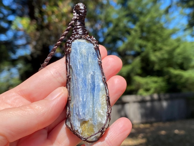 May include: A blue kyanite pendant necklace. The raw, elongated crystal is wrapped in brown wire and suspended from a matching cord. The stone has streaks of blue and yellow hues. The background is blurred with green foliage.
